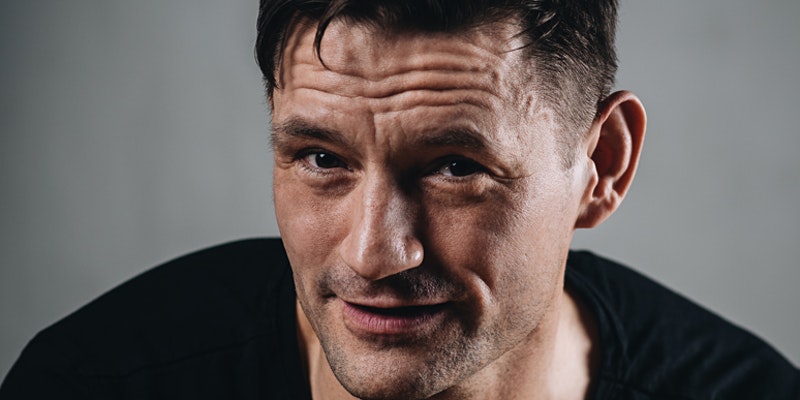 Gunnar Söderström porträttbild. Han hr kort mörkt snaggat hår, svart tröja och tittar snett in i kameran. Han har kort skäggstubb och har grå bakgrund.