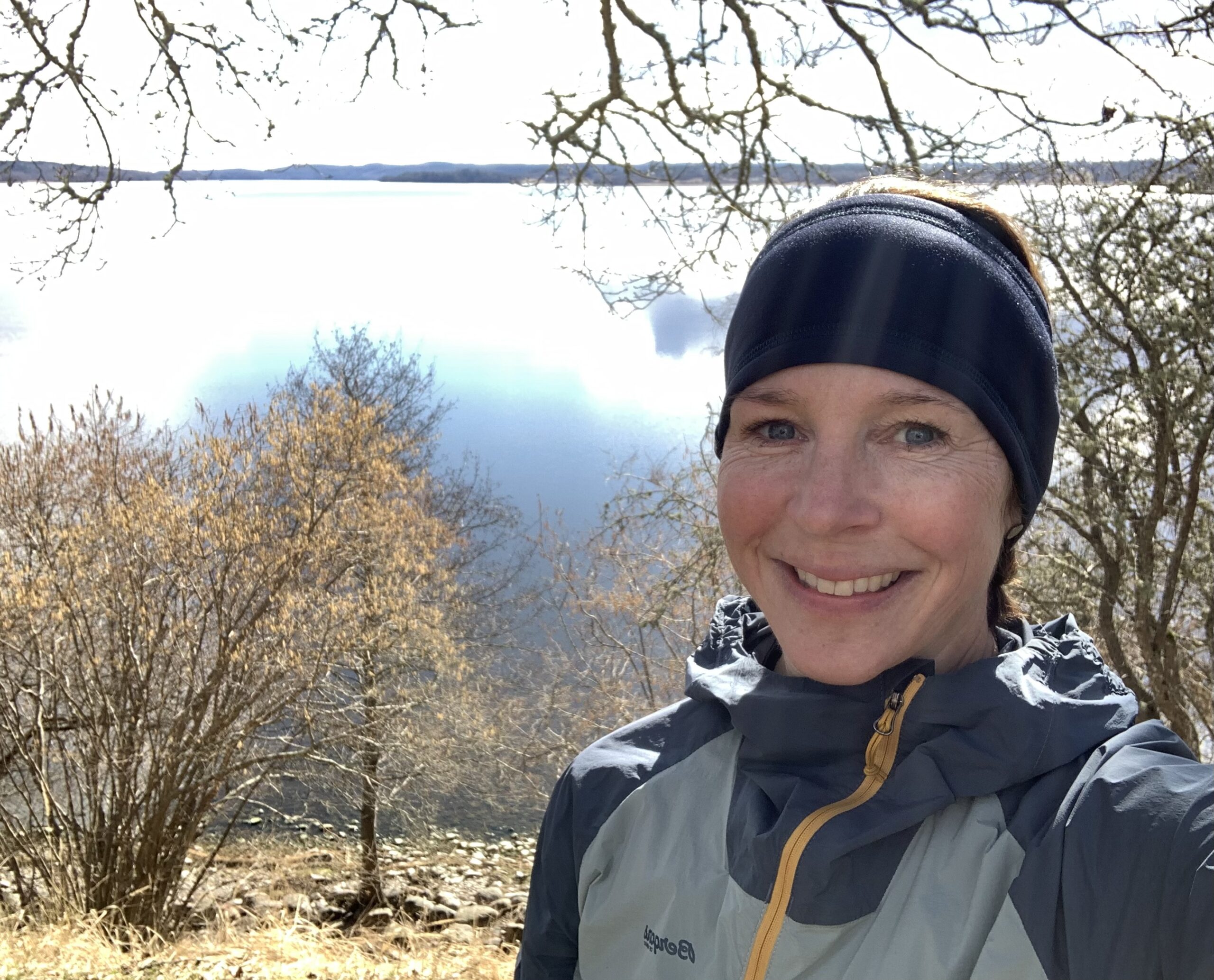 Profilbild Sofia Hamrå. Hon står utomhus med gråblå träningskläder och ler. I bakgrunden syns en sjö och träd.
