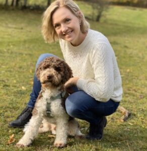 Kristina Dellholm med sin hund. Kristina har axellångt blont hår, vit långärmad tröja och sitter på en gräsmatta intill sin hund, en brunvit pudel.