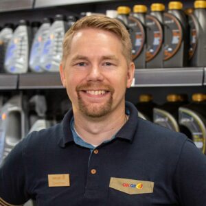 Jonathan står i sin butik med flaskor på en hylla bakom sig. Han har kort ljust hår, skägg och mustasch. Han är klädd i en mörk arbetsklädsel och ser glad ut.