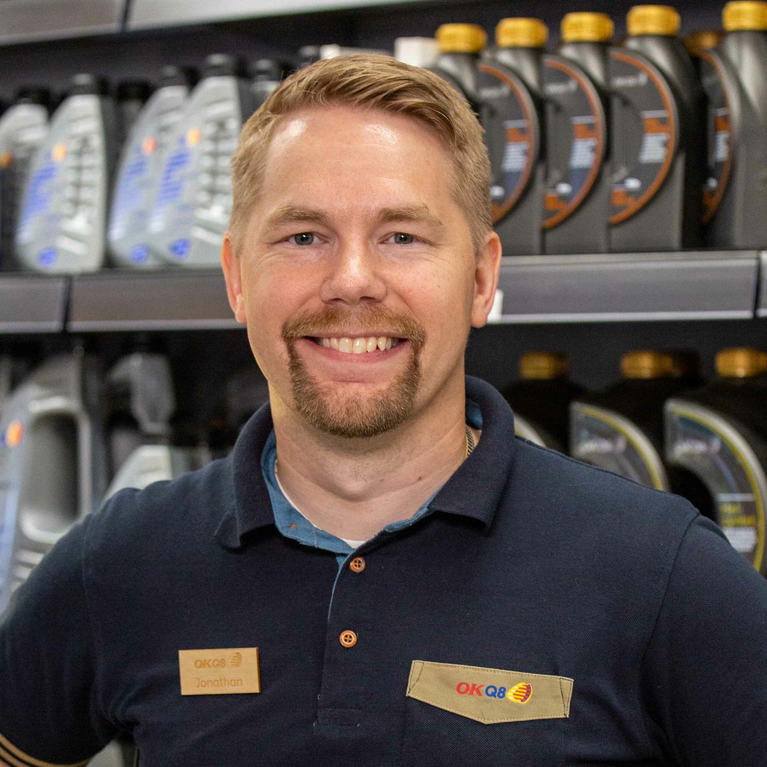 Jonathan står i sin butik med flaskor på en hylla bakom sig. Han har kort ljust hår, skägg och mustasch. Han är klädd i en mörk arbetsklädsel och ser glad ut.