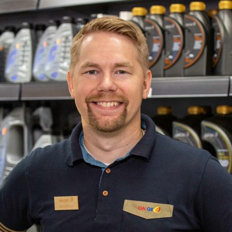 Jonathan står i sin butik med flaskor på en hylla bakom sig. Han har kort ljust hår, skägg och mustasch. Han är klädd i en mörk arbetsklädsel och ser glad ut.