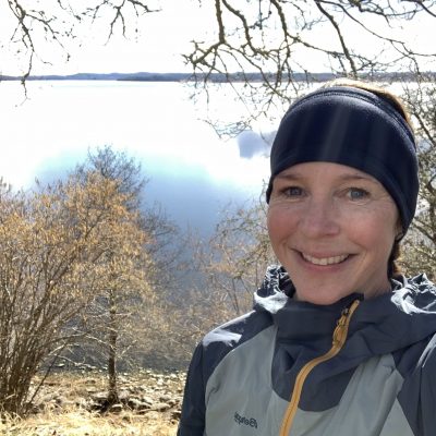 Profilbild Sofia Hamrå. Hon står utomhus med gråblå träningskläder och ler. I bakgrunden syns en sjö och träd.
