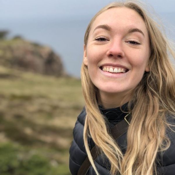 Tea Svensson utomhus, profilbild. Hon har ljusblont långt hår och ler. Berg och sjö i bakgrunden.