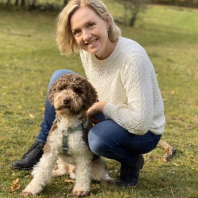 Kristina Dellholm med sin hund. Kristina har axellångt blont hår, vit långärmad tröja och sitter på en gräsmatta intill sin hund, en brunvit pudel.
